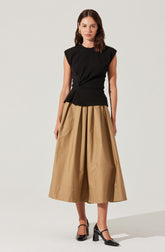 A woman is wearing a sleeveless black top with structured shoulder details and a high neckline, paired with a long, wide-leg beige skirt that reaches her ankles. She has on black shiny heels with ankle straps.